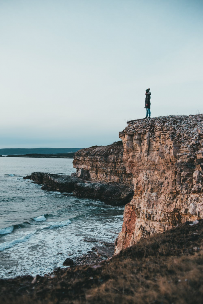 Woman standing on side of a rocky cliff, looking out at the ocean.