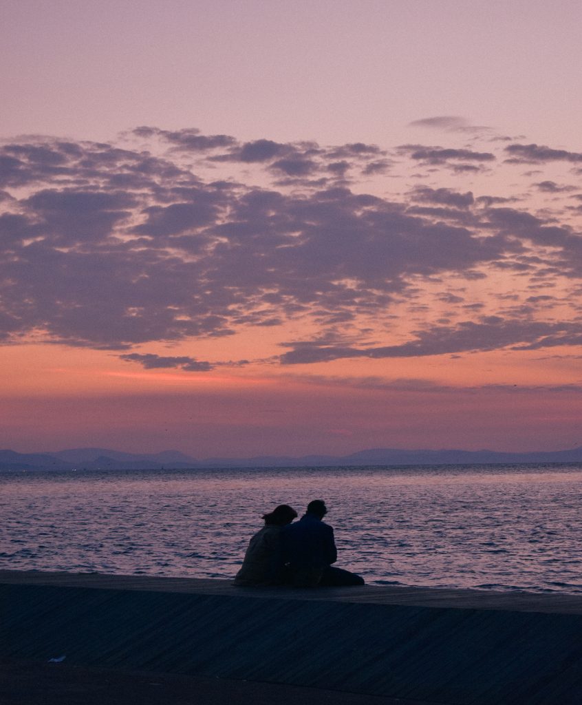 Silhouette of a couple sitting in front of a calm ocean scape and cloudy sunset sky.